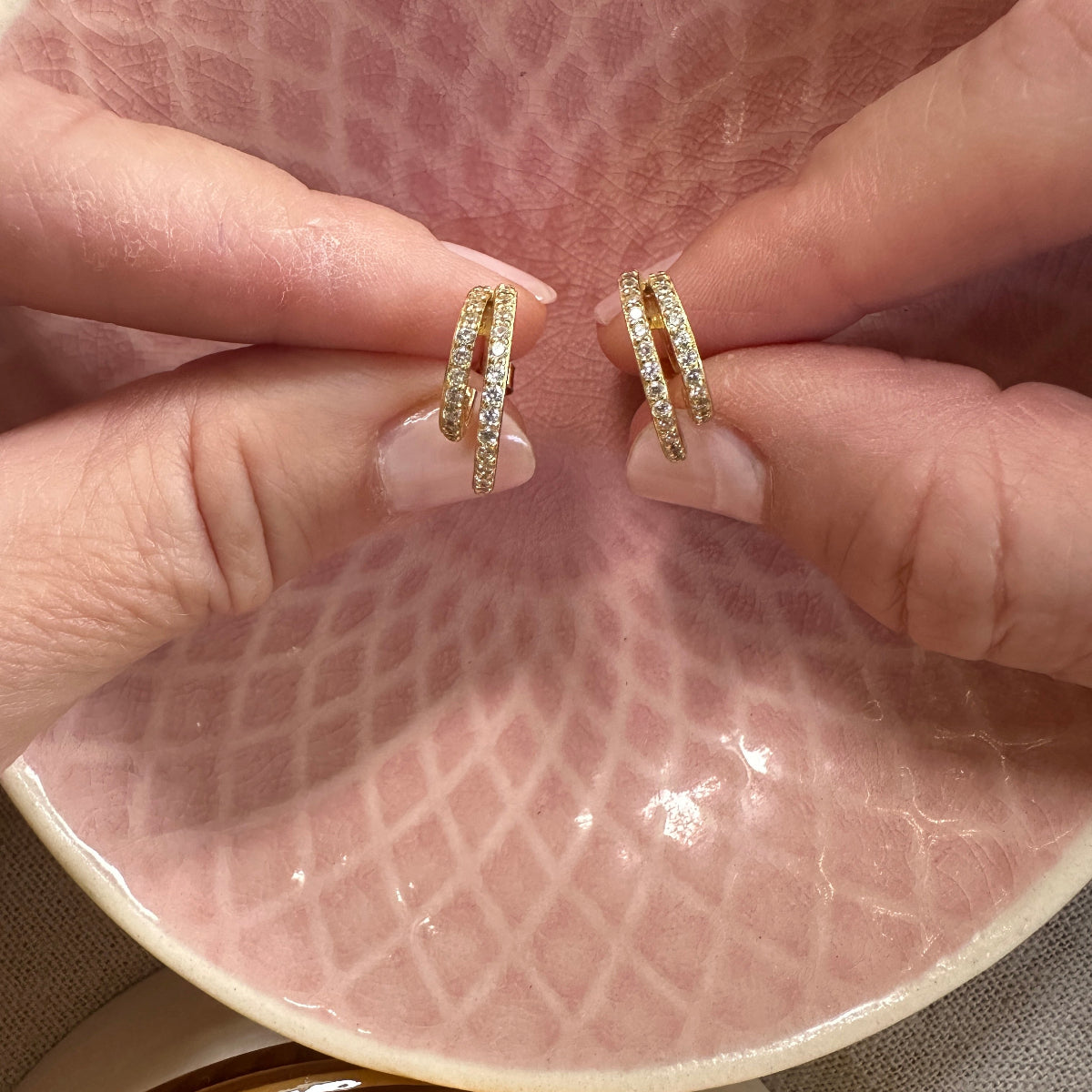Gold hoop earrings held between fingers on a textured pink surface