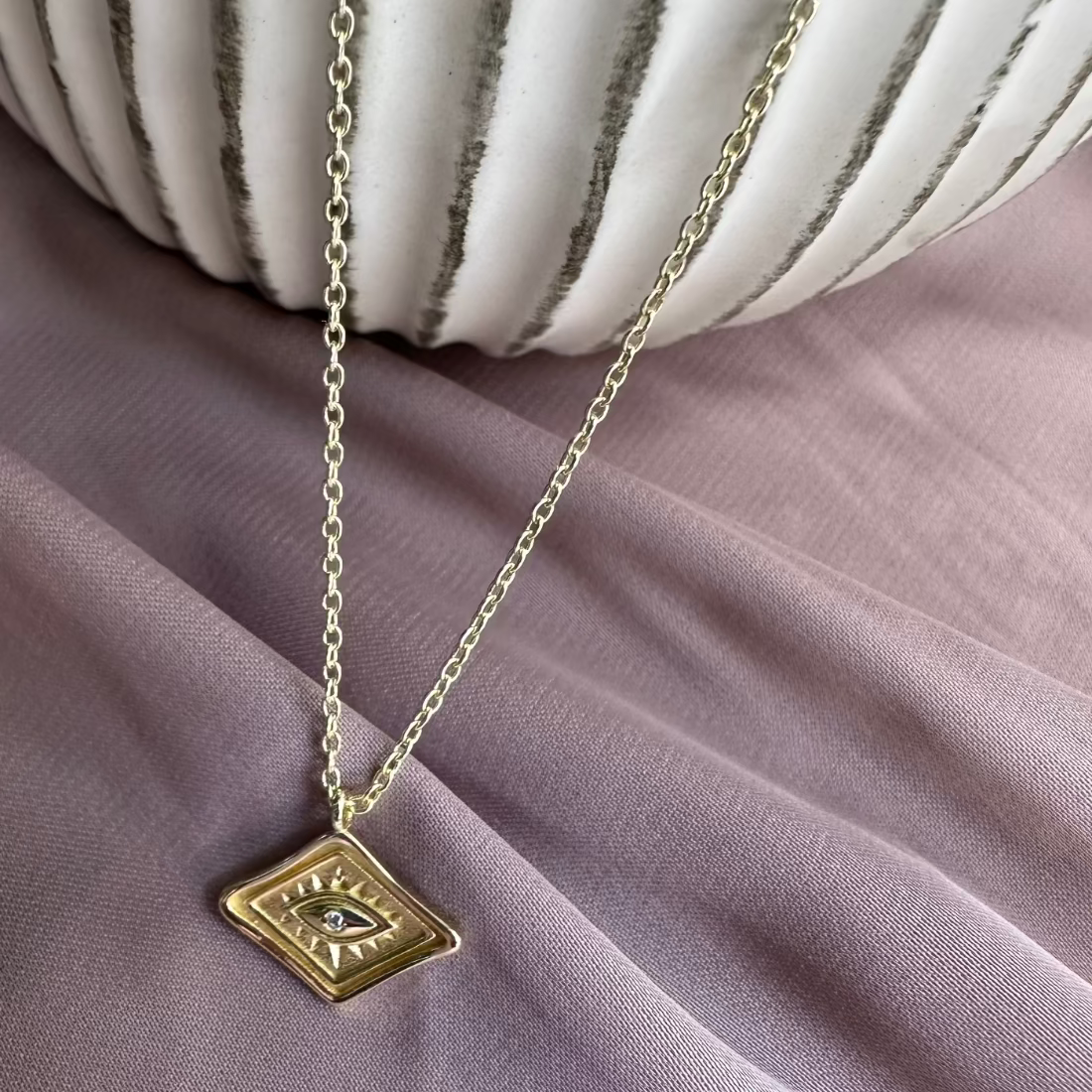 gold diamond shaped pendant with a cubic zirconia gemstone with eye style detail. laid on pink ground
