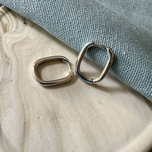 A pair of silver oval hoop earrings placed on a textured surface next to a blue fabric.