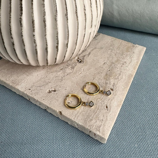 A pair of gold hoop earrings with a gemstone charm, displayed on a textured surface next to a white, abstract-patterned object.