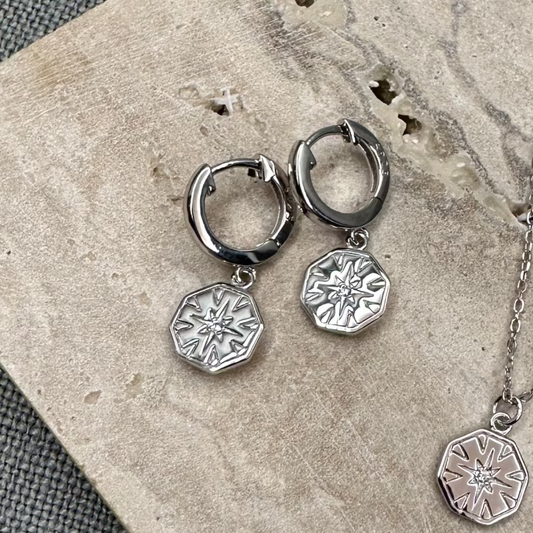 A pair of silver-colored hexagon charm huggie earrings with a geometric design, displayed on a textured background.