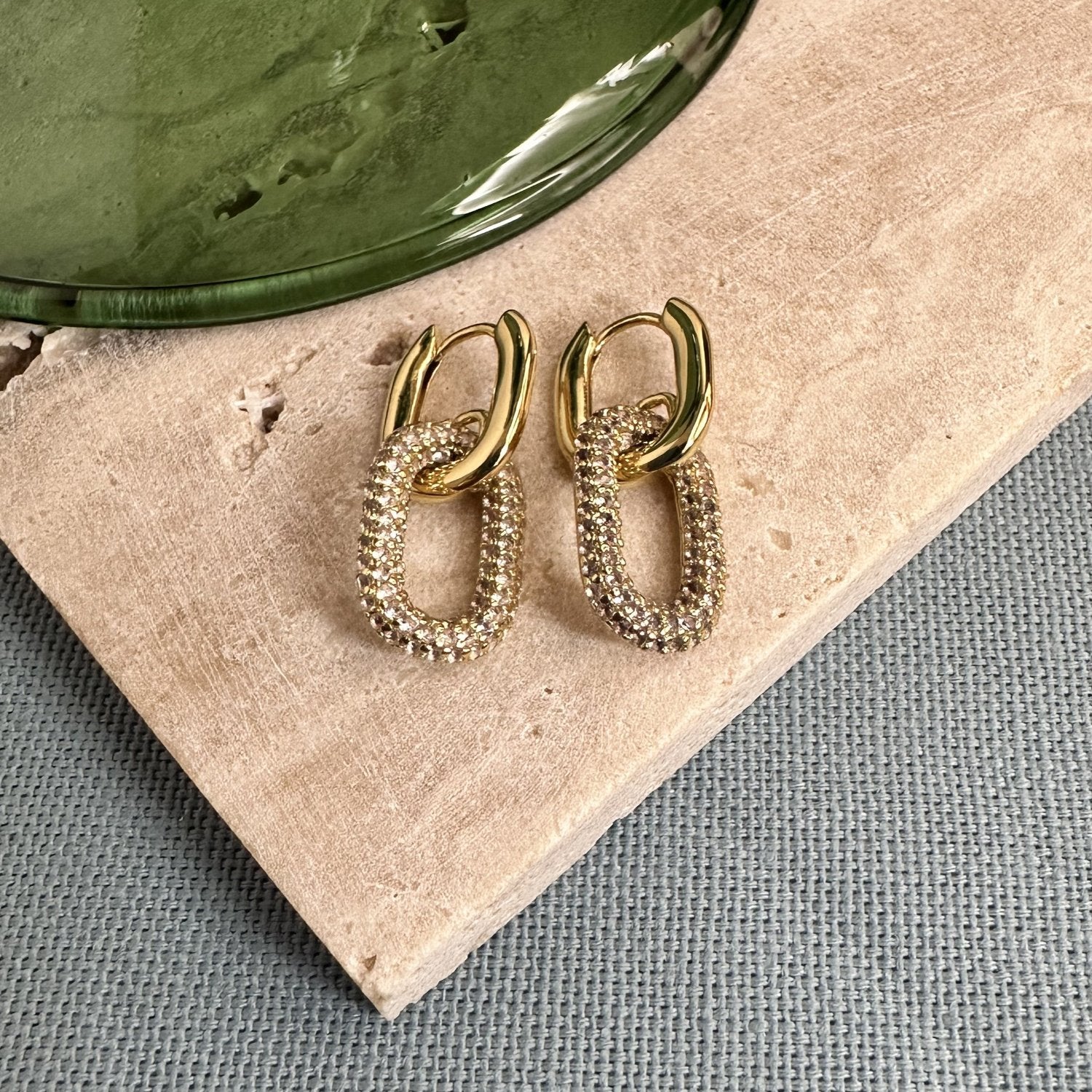 Gold oval-shaped huggie earrings with a pave gemstone charm on a wooden surface.