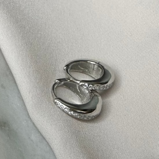 A pair of silver huggie hoop earrings with a smooth, domed design, displayed on a silky surface.