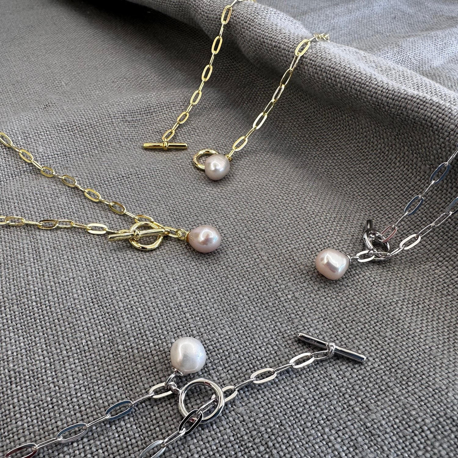 A collection of jewellery laid out on a textured surface, including a gold paperclip chain bracelet with a pearl charm and a matching necklace.