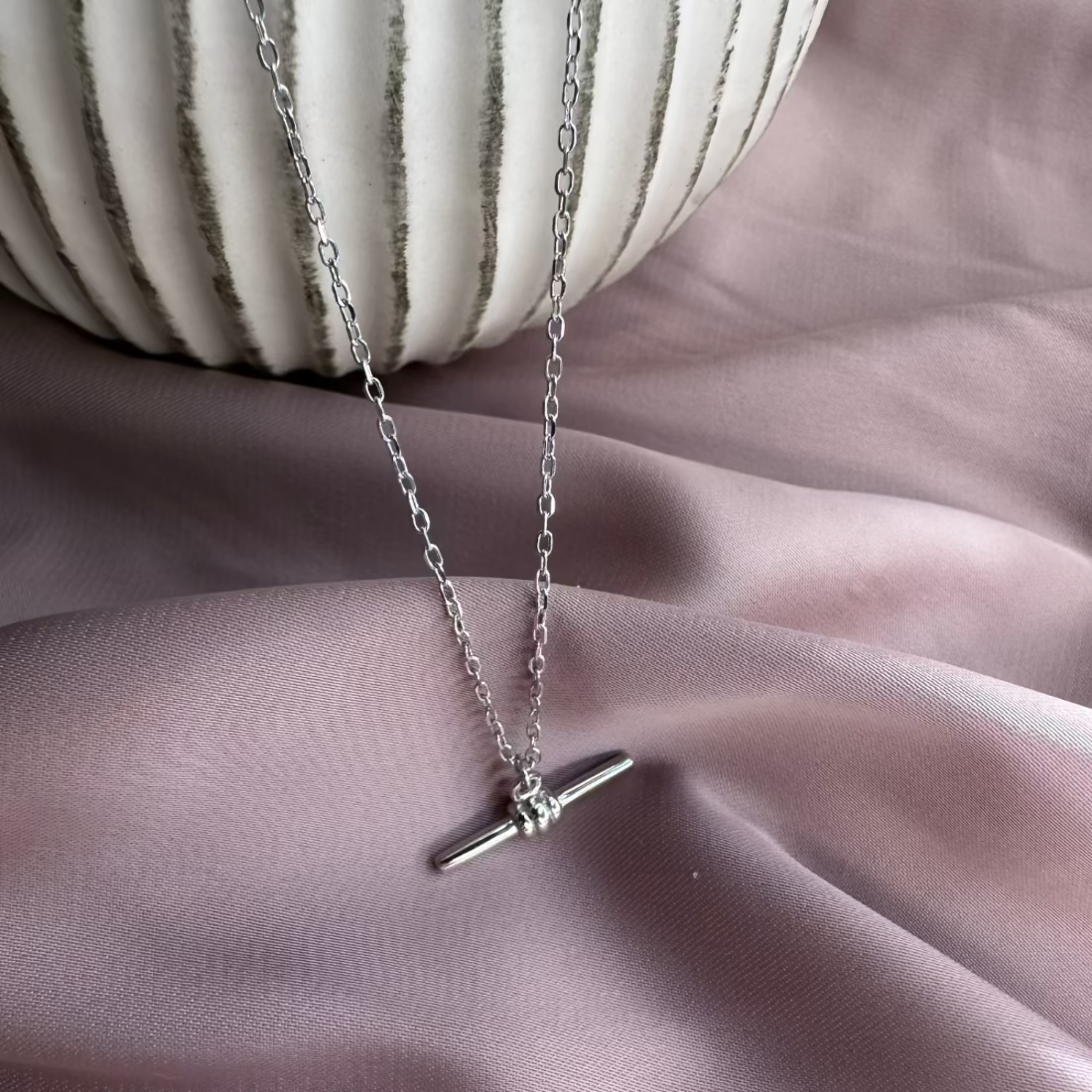 a delicate t-bar pendant on a fine chain laid on pink fabric and cermaiv vase