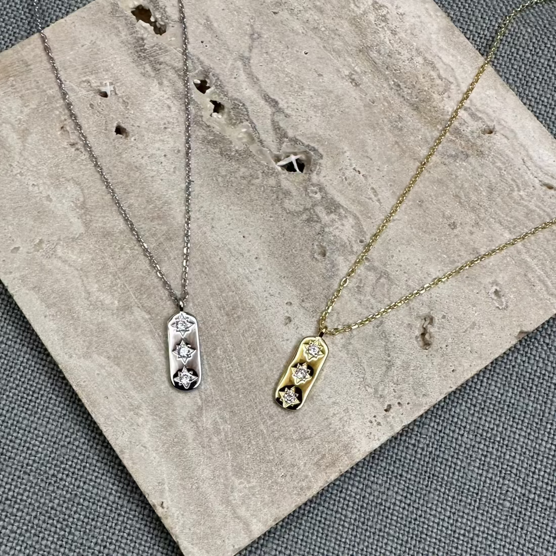 A gold and a silver necklace with a geometric pendant featuring three sparkling white cubic zirconia stones, displayed on a textured grey stone.