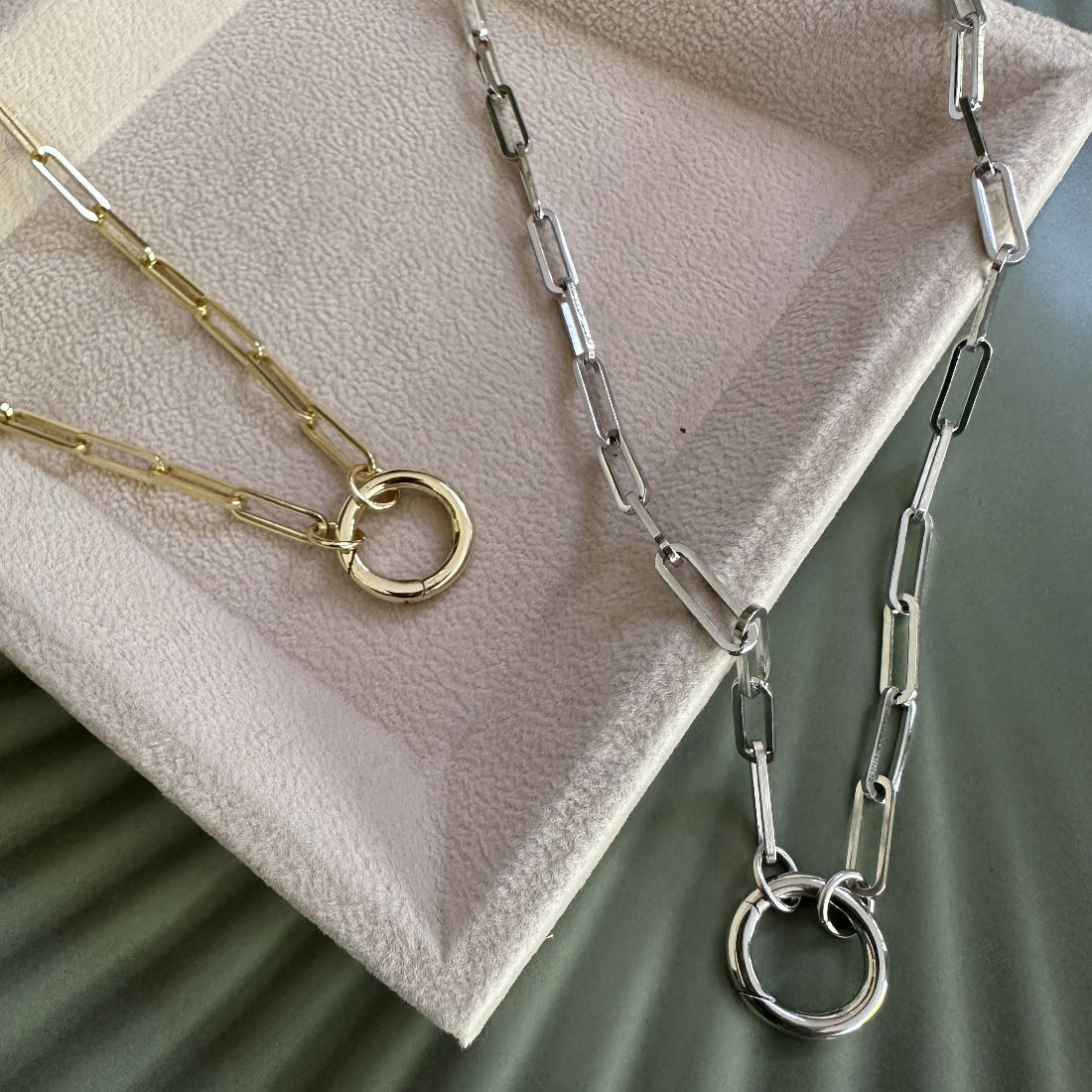 Two paperclip statement necklaces in Silver and Gold with a charm carrier laid on neutral tray