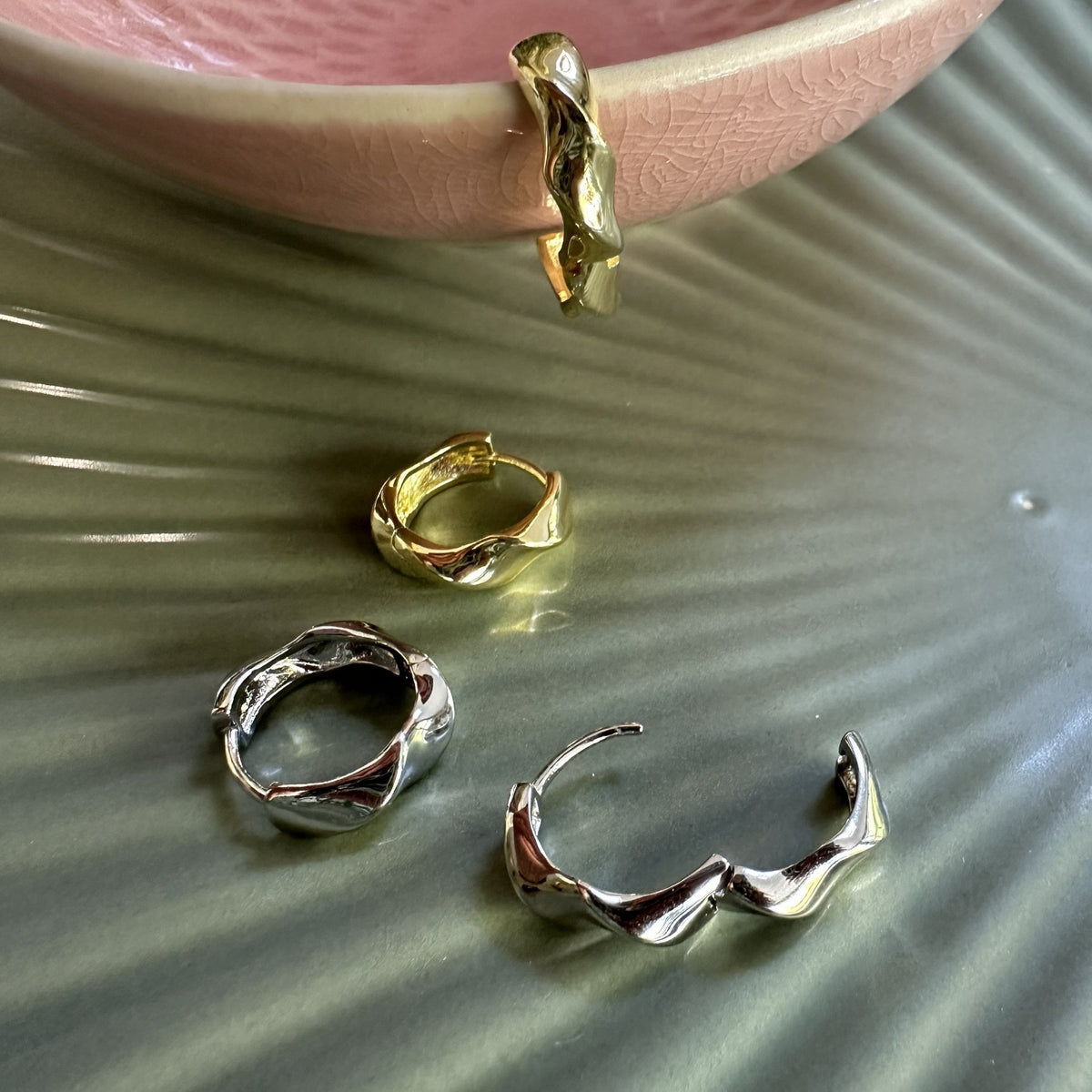 Gold and silver chunky modern hoop earrings on a textured ceramic surface with a pink dish in the background.