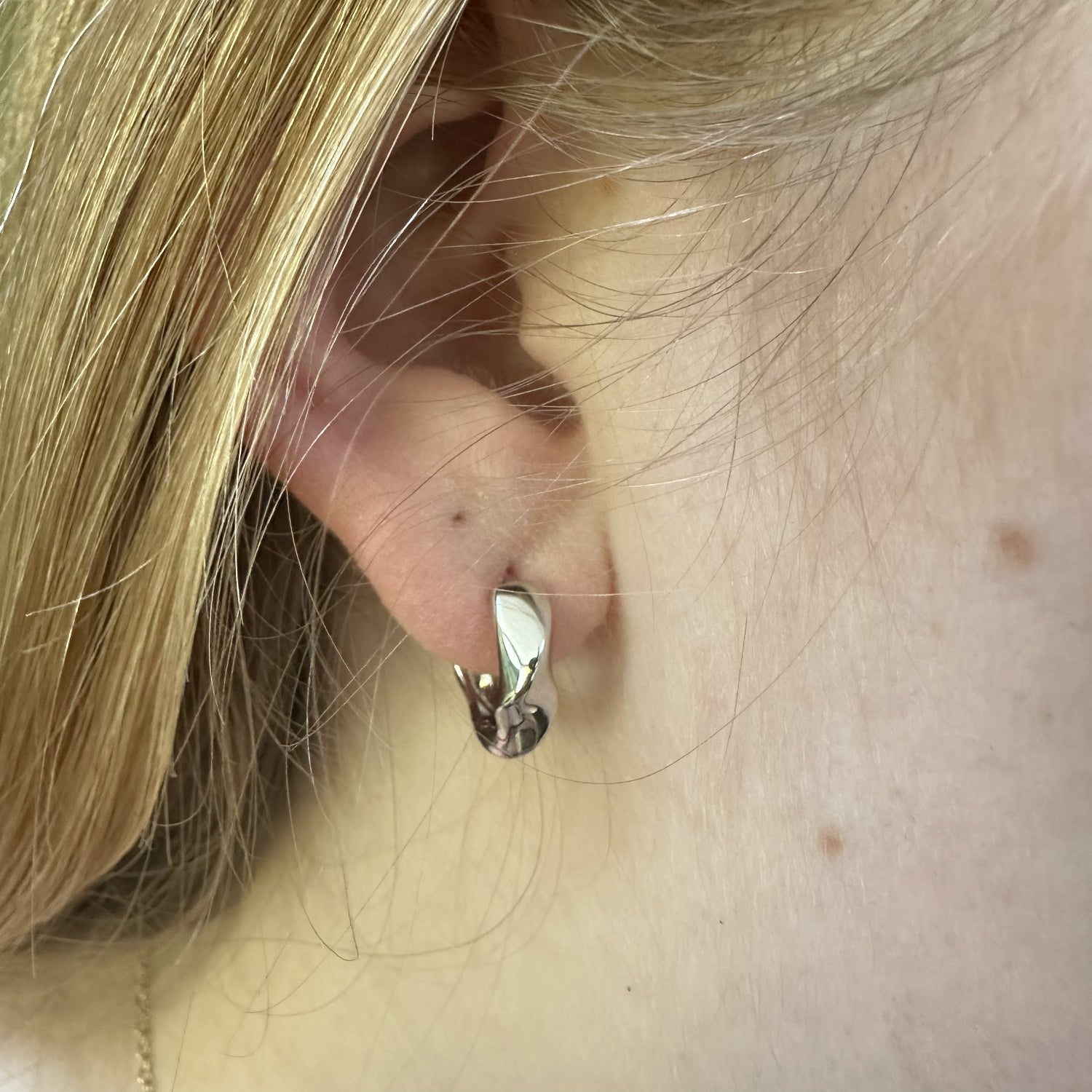Close-up of an ear wearing a medium sized statament chunky silver earring with blonde hair partially covering it.