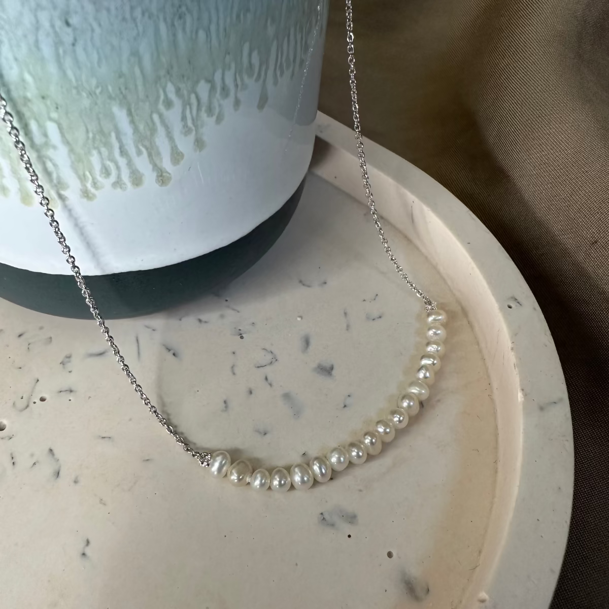 A white freshwater pearl necklace with an adjustable slider is displayed on a silver chain, presented on a neutral-toned surface.