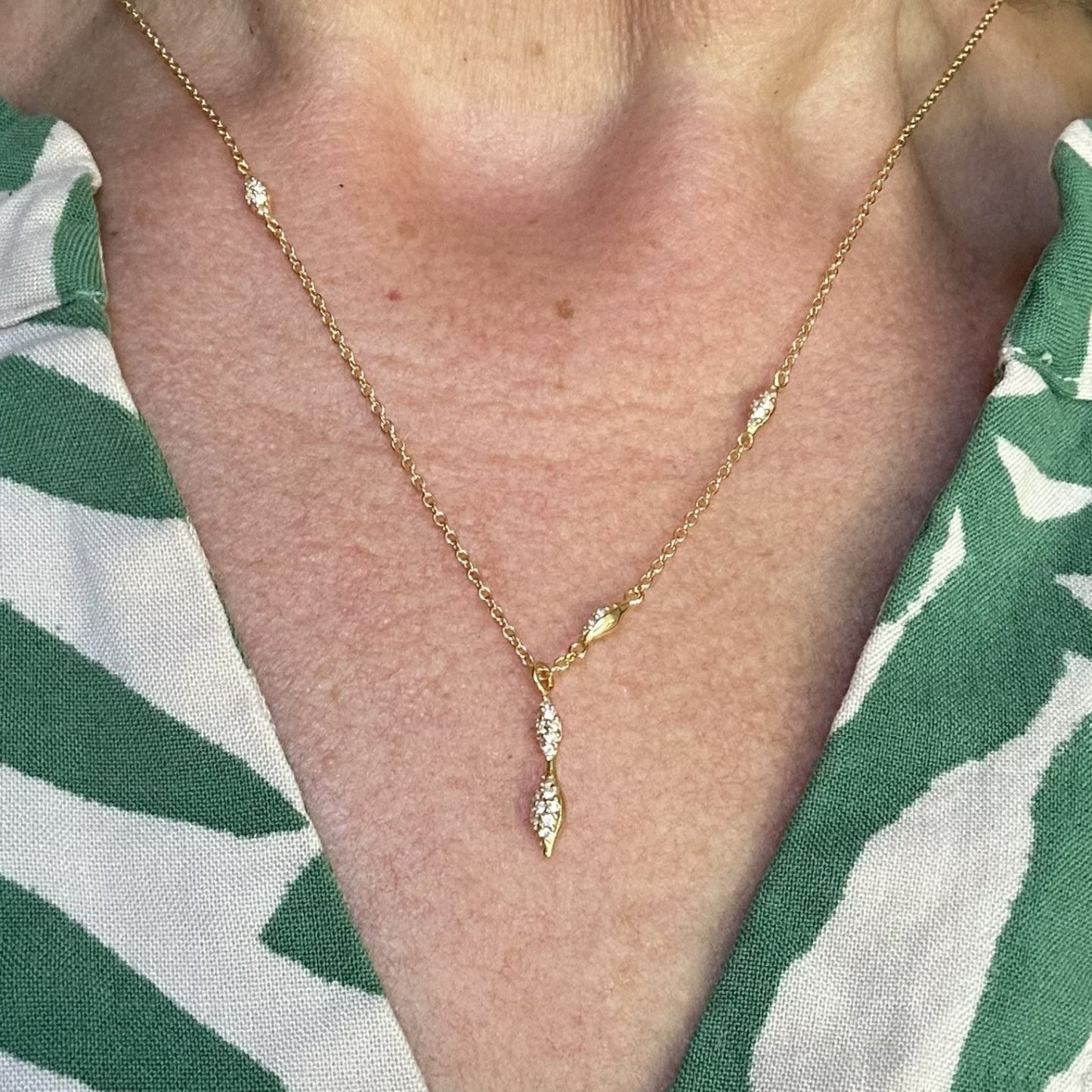 Gold necklace with a modern sparkly drop pendant on a person wearing a green and white patterned shirt.