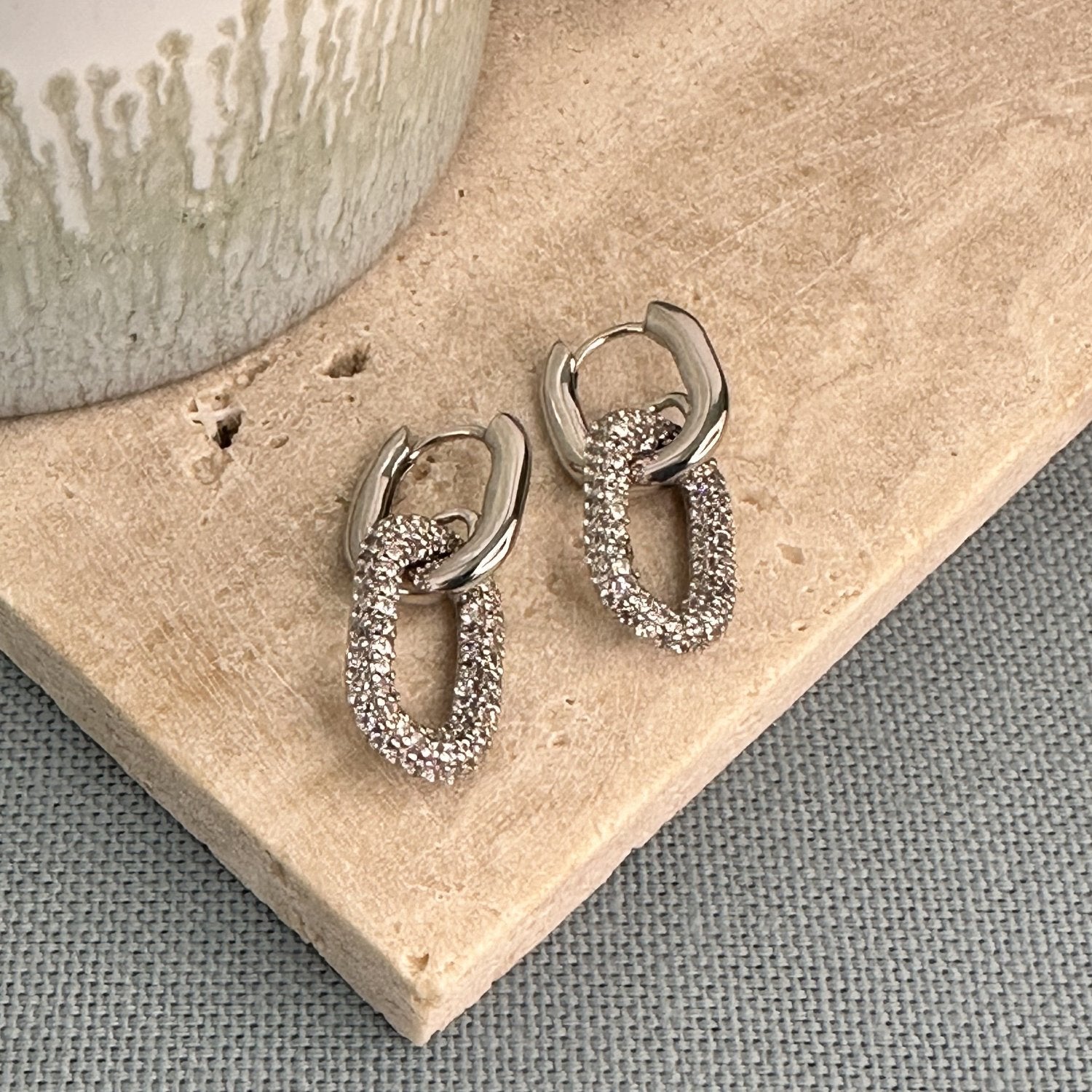 Silver oval-shaped huggie earrings with a pave gemstone charm on a wooden surface.