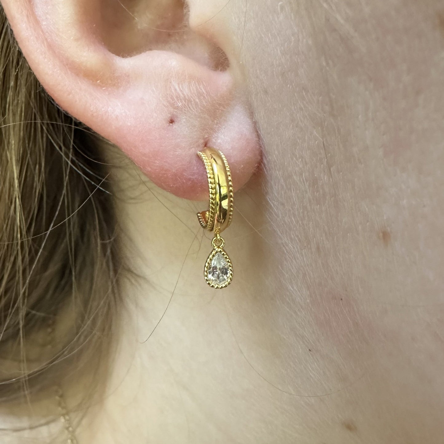 Close-up of a gold hoop earring with a teardrop-shaped pendant worn by a person.