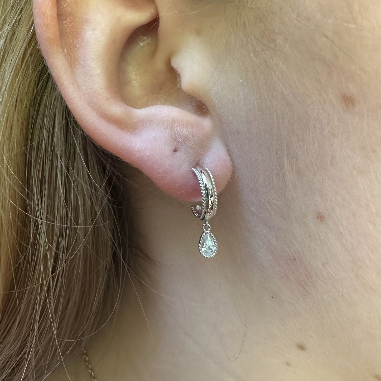 Close-up of an ear wearing a silver hoop earring with a teardrop-shaped pendant.