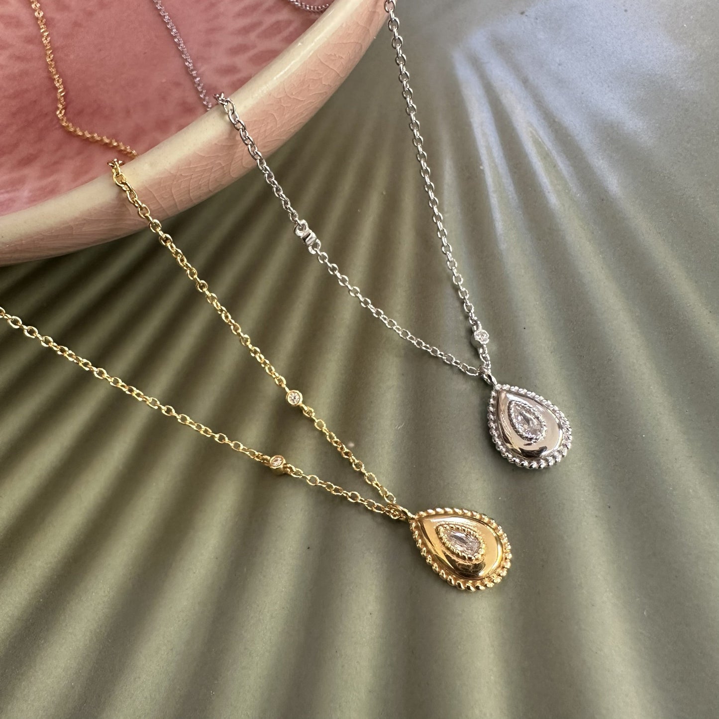 Two gold necklaces with teardrop pendants on a textured surface.