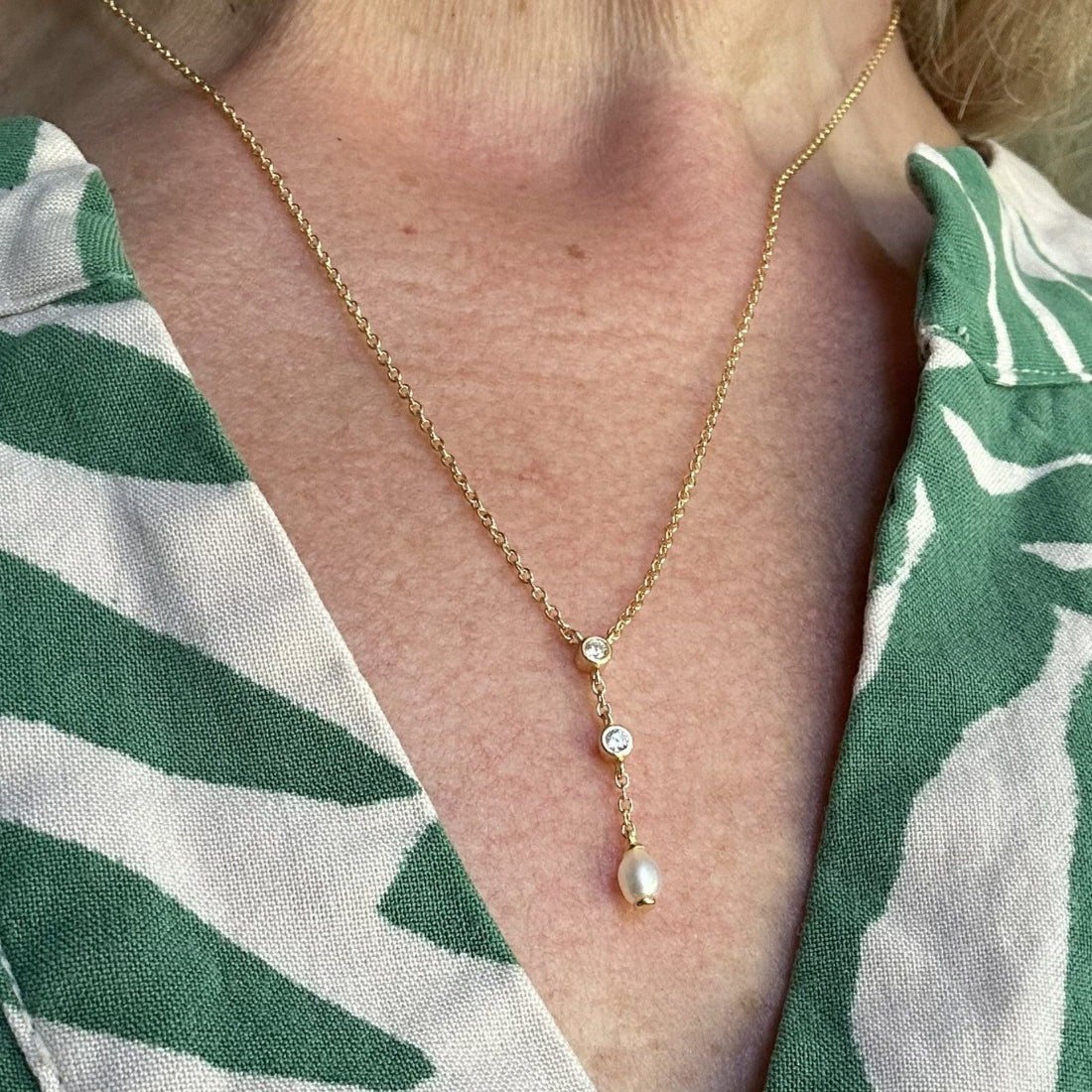 Gold necklace with a natural freshwater pearl and sparkle gemstone pendant worn by a person in a green and white patterned shirt.