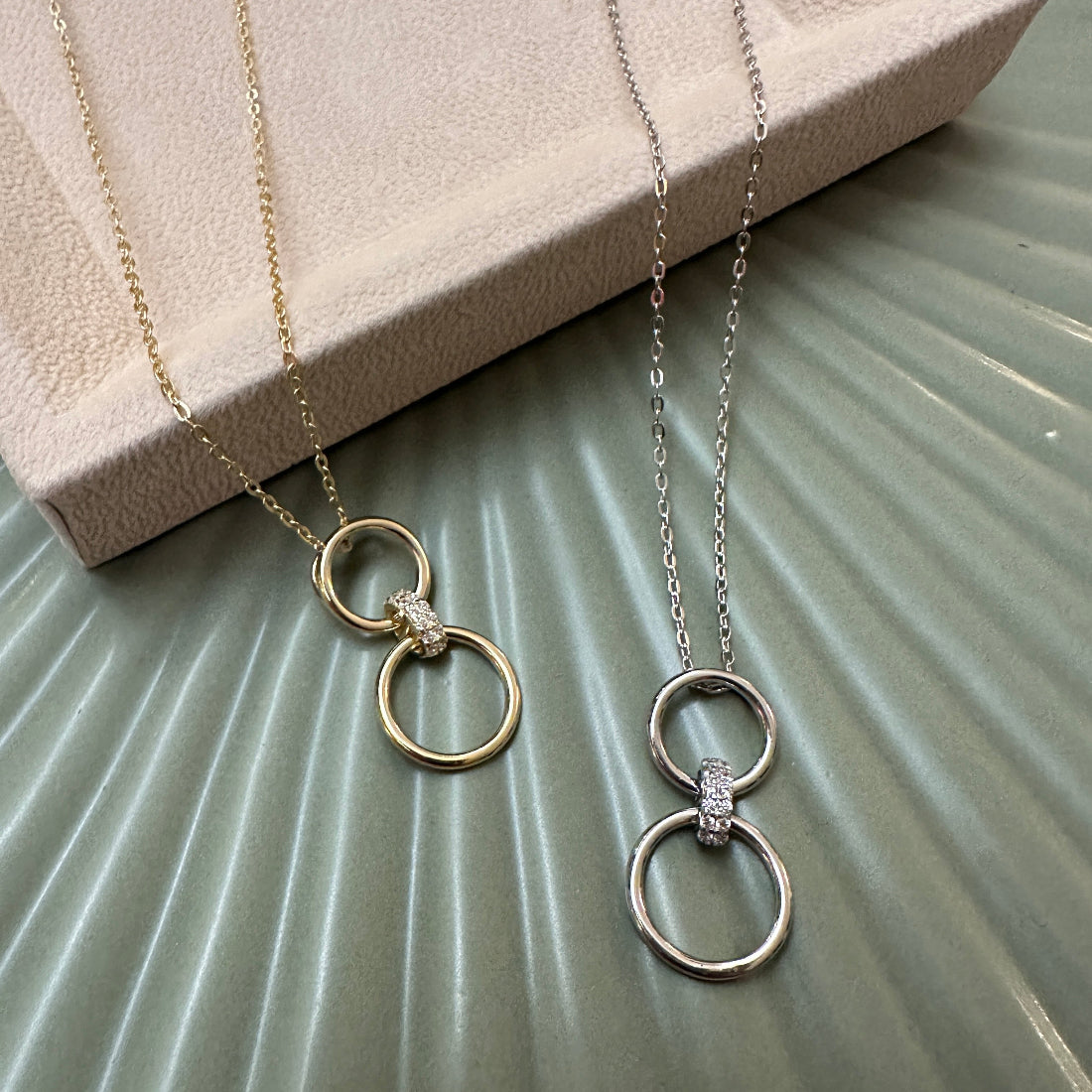 A silver and gold pendant necklace both with double circle connected with a sparkling interlocking ring to bound the rings together, on green ground