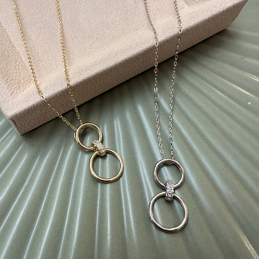 A silver and gold pendant necklace both with double circle connected with a sparkling interlocking ring to bound the rings together, on green ground