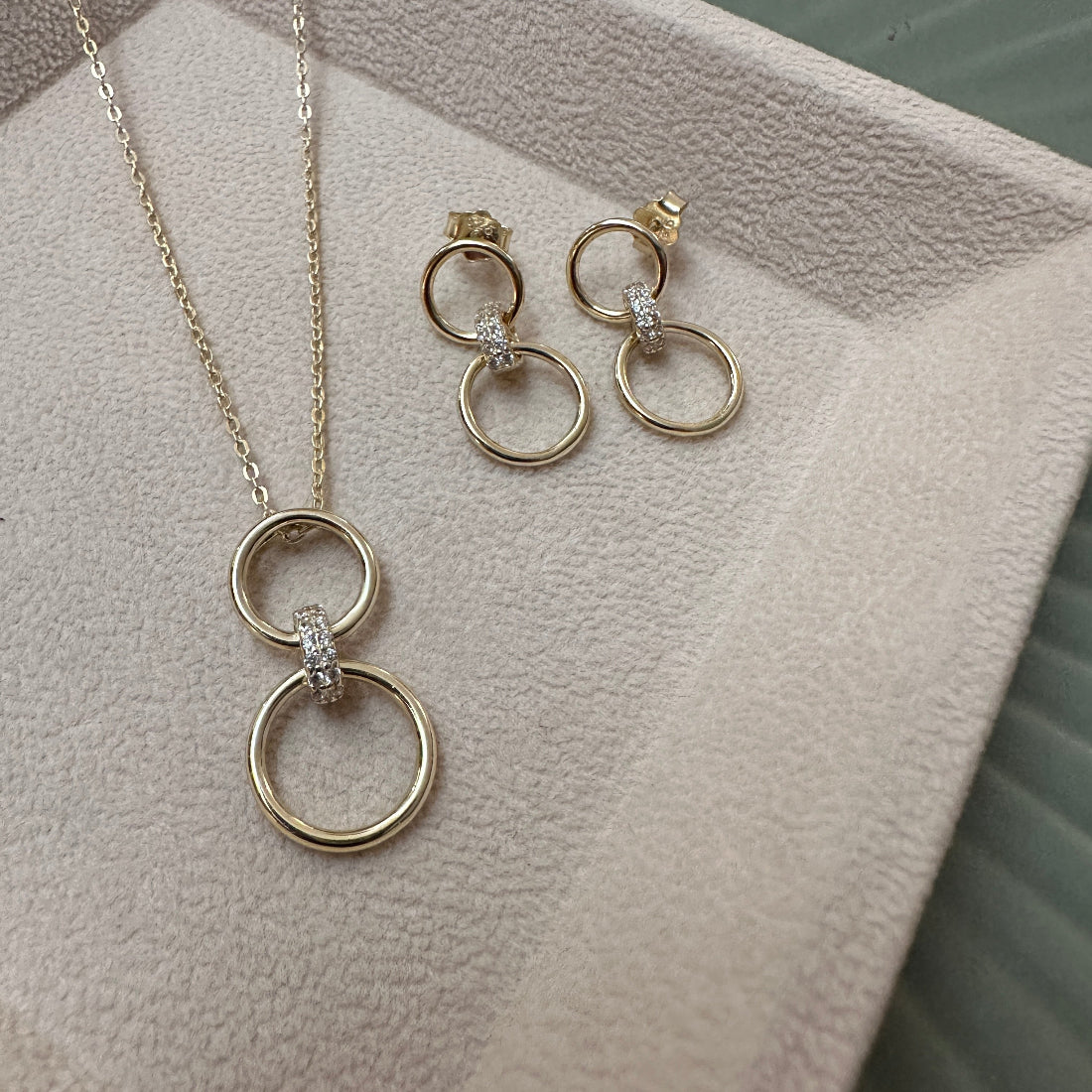 Gold necklace and earrings featurings a drop pendant with 2 ciclres connected with a sparkly link set on a textured surface