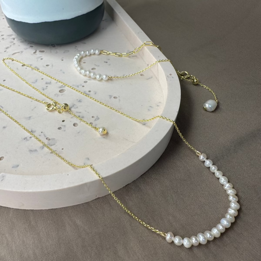 A white freshwater pearl necklace with an adjustable slider is displayed on a gold chain, accompanied by a matching bracelet and a pearl drop earring, presented on a neutral-toned surface.
