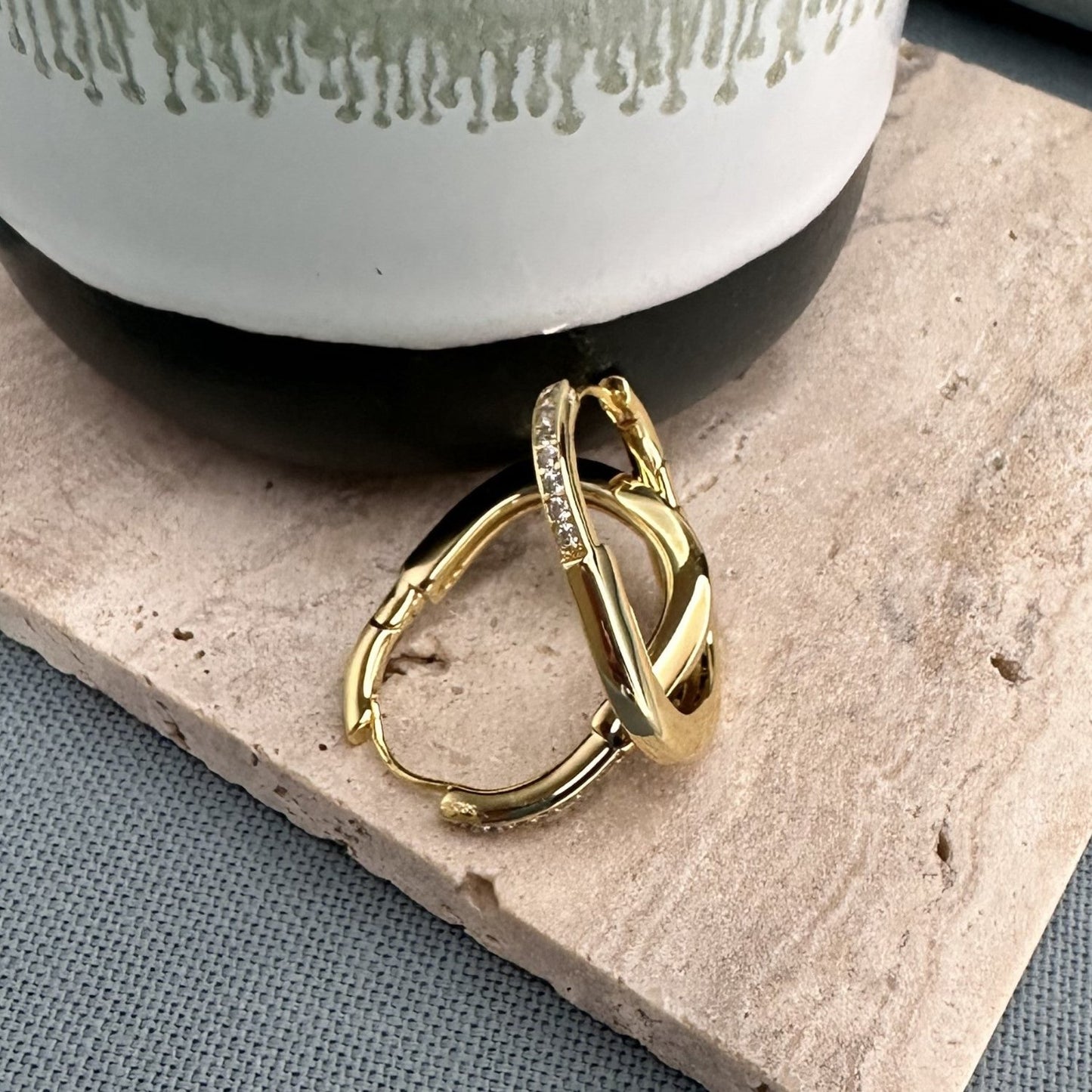 A pair of large oval-shaped gold hoop earrings with a secure back hinge fastening, placed on a textured surface.