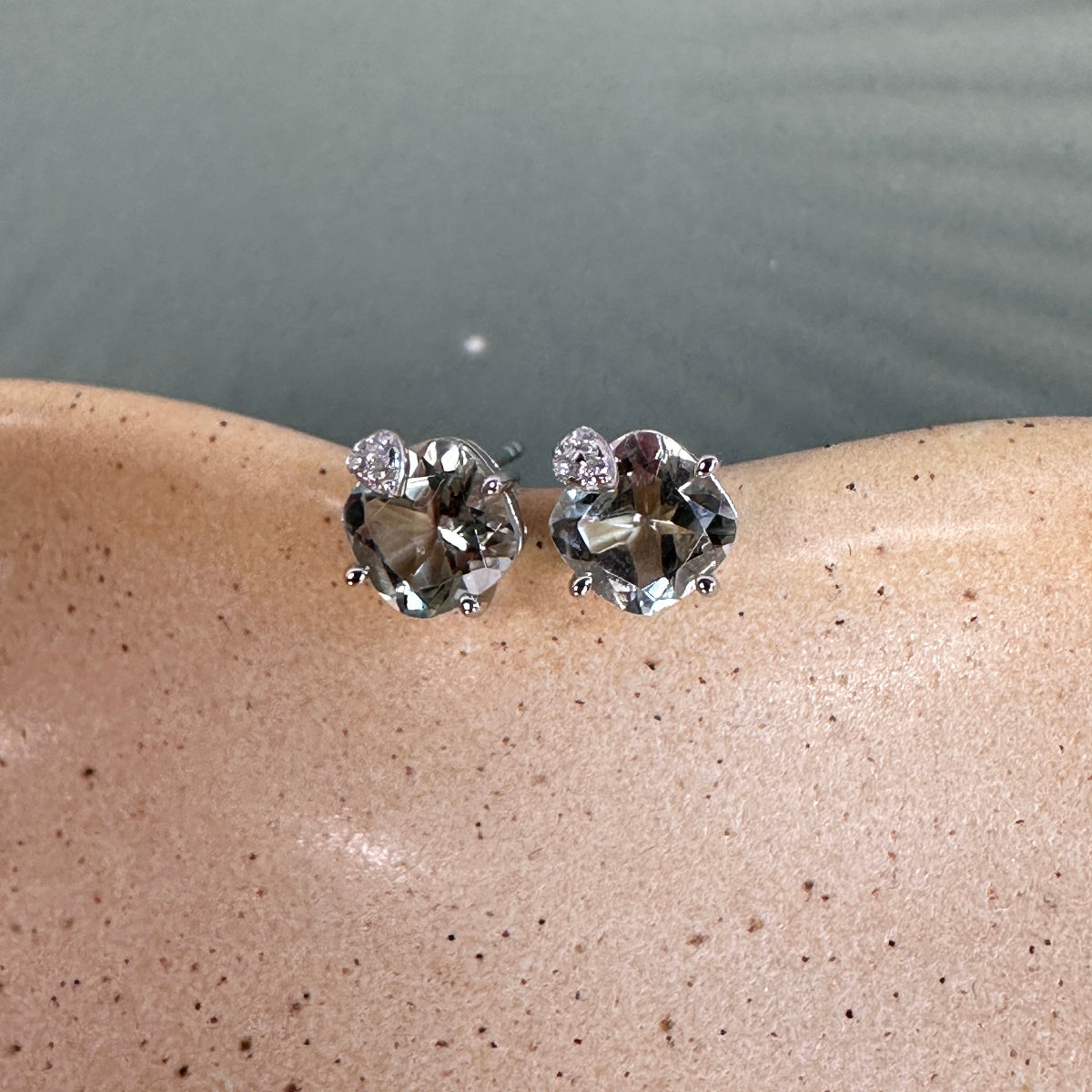 Pair of silver stud earrings with cushion cut green amethyst gemstones on a beige surface with a blurred background