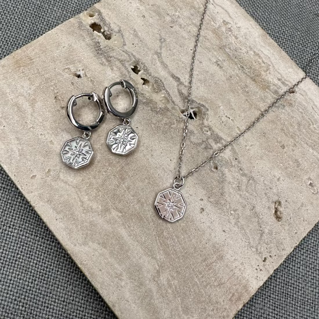 A pair of silver hexagon charm huggie earrings and matching necklace with a geometric design, displayed on a textured background.