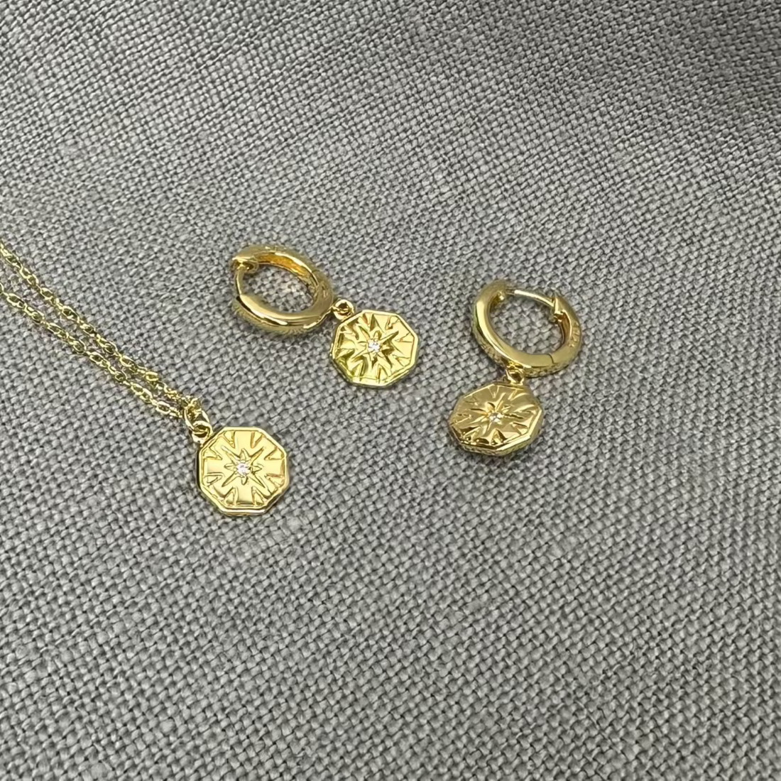 A pair of gold hexagon charm huggie earrings with a geometric design, displayed on a textured background