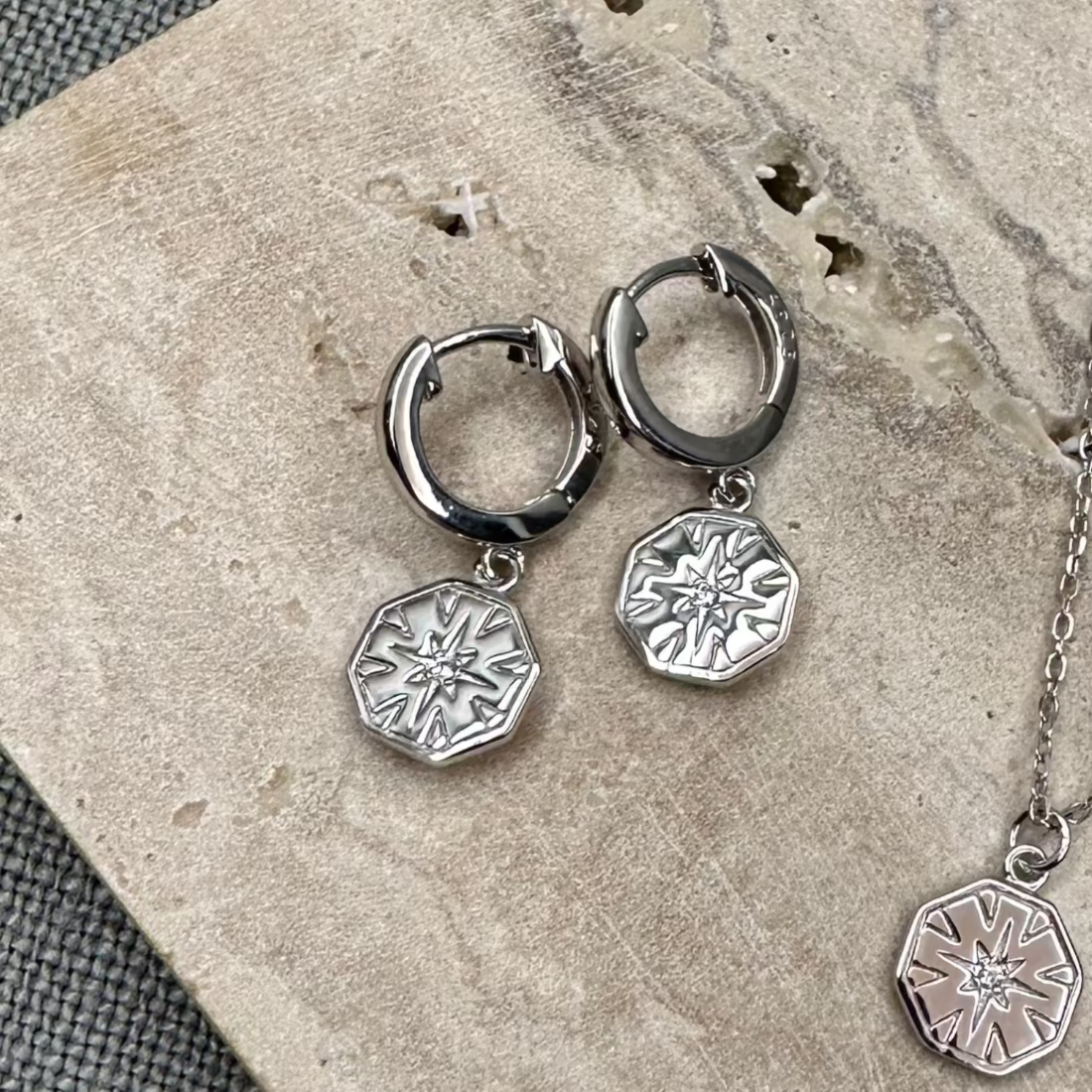 A pair of silver-colored hexagon charm huggie earrings with a geometric design, displayed on a textured background.