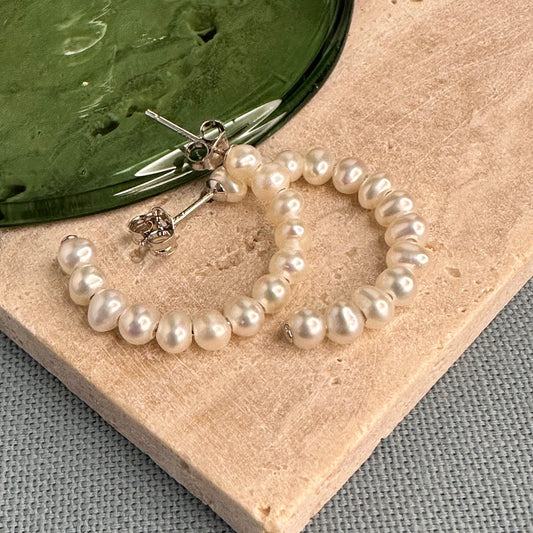 A pair of large pearl hoop earrings displayed on a wooden surface with a blurred green background.