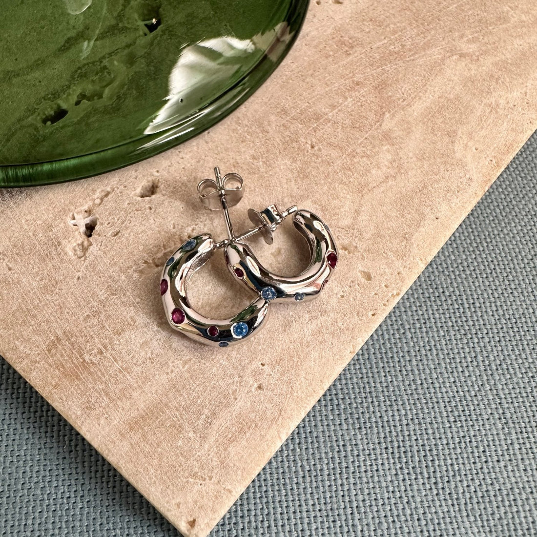 mini sized silver hoops in an organic shape set with multicoloured gemstones laid on a tile