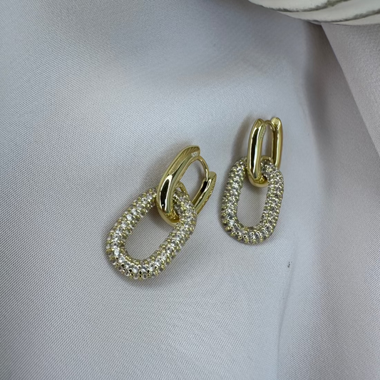 A pair of gold huggie earrings with pave gemstone charms on a white fabric surface.