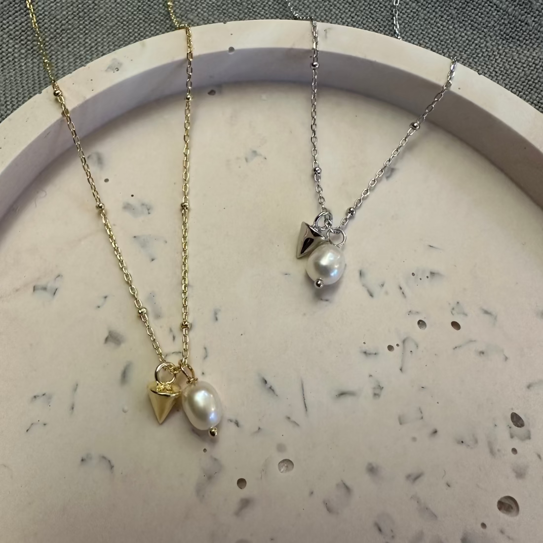 two pendant necklaces in gold and silver, each featuring a freshwater natural pearl and a metal gone shaped charm laid on a marble tray