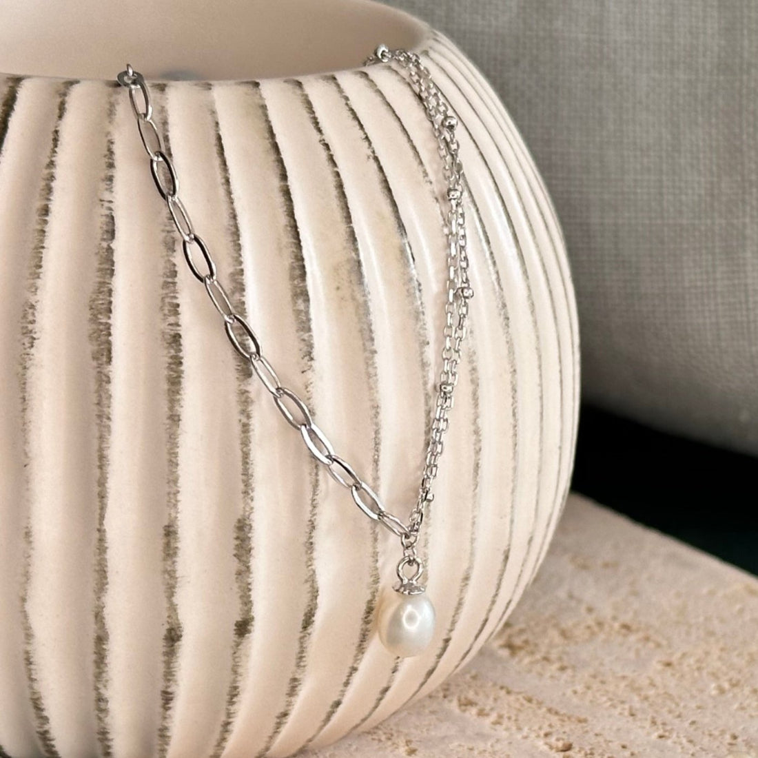 A sterling silver necklace with a double satellite chain and a paperclip chain, featuring a pearl drop pendant, displayed on a textured surface.