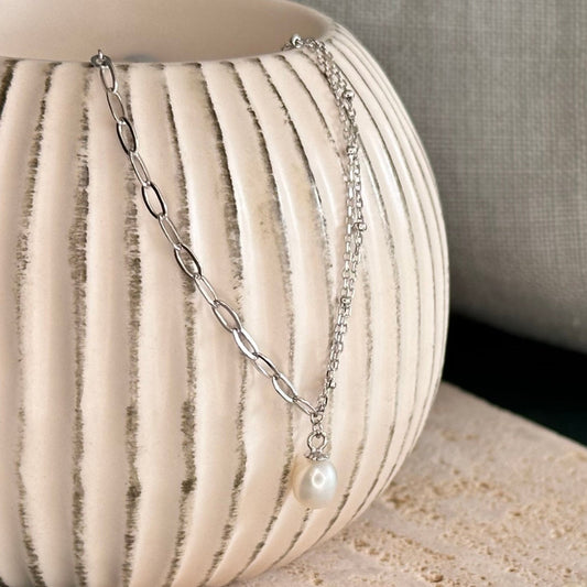 A sterling silver necklace with a double satellite chain and a paperclip chain, featuring a pearl drop pendant, displayed on a textured surface.