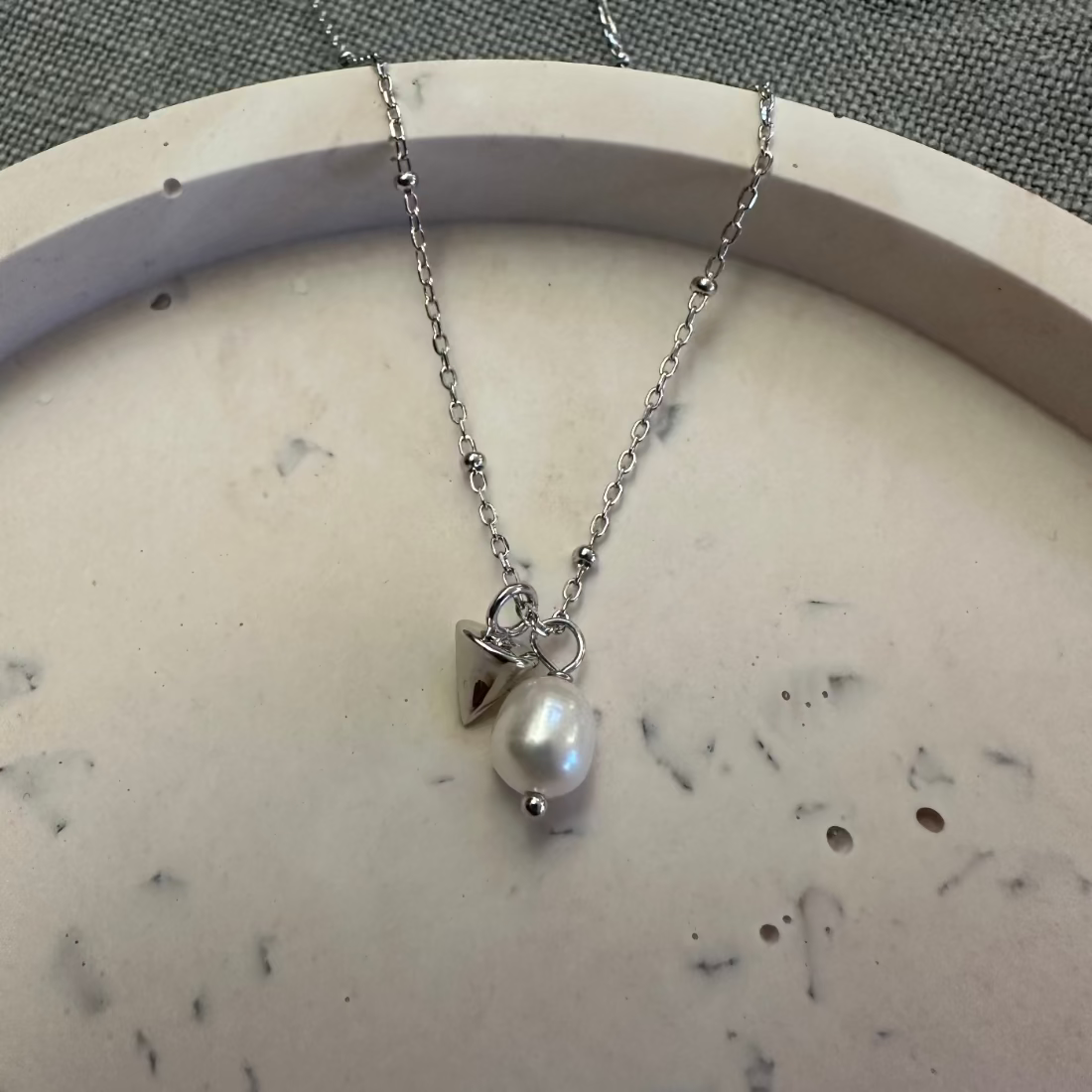 a modern freshwater pearl necklace on a trending ball chain with a silver cone charm laid on marble tray
