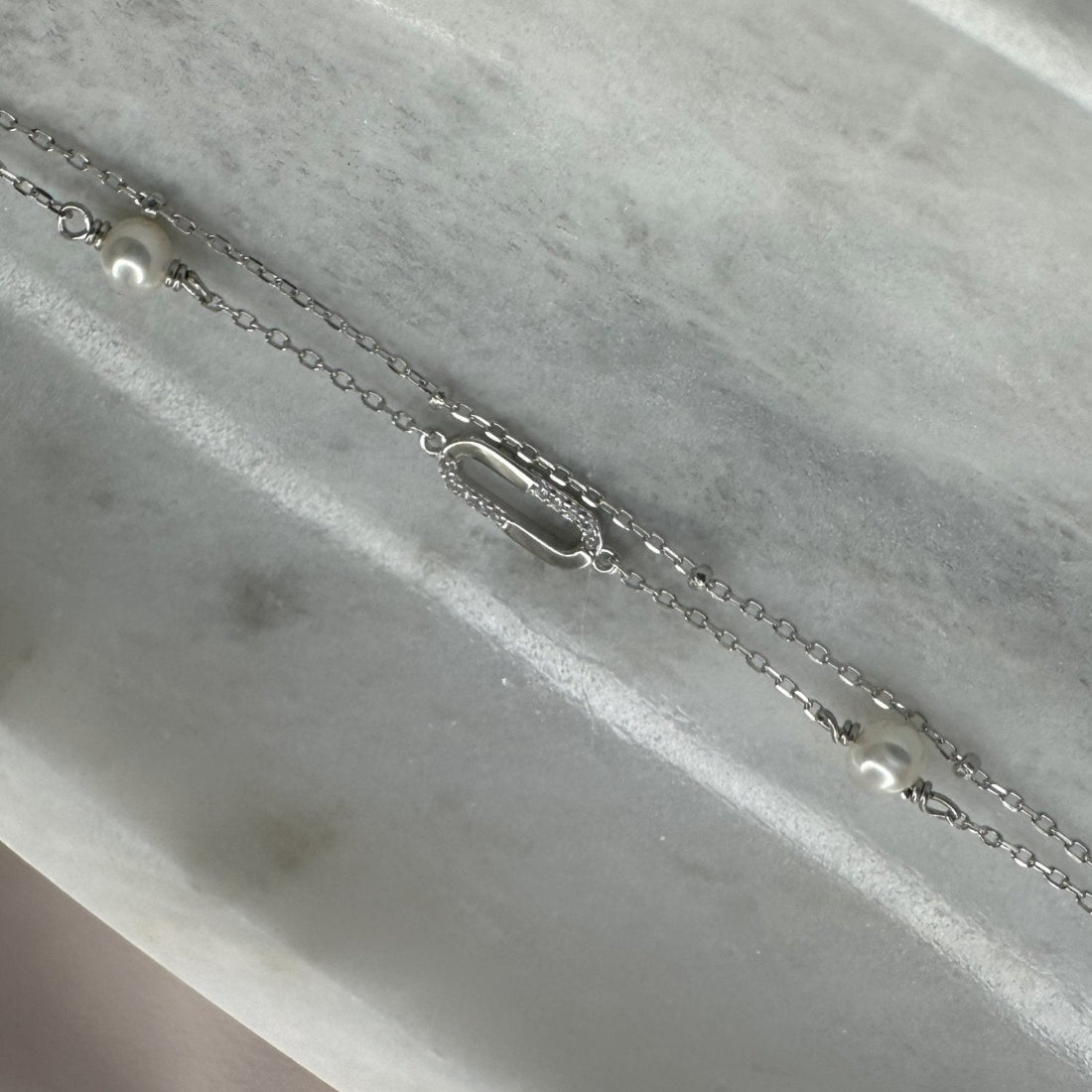 a close up of a double chain with pearl and oval charm with gemstones encrusted on the top. laid on a marble dish