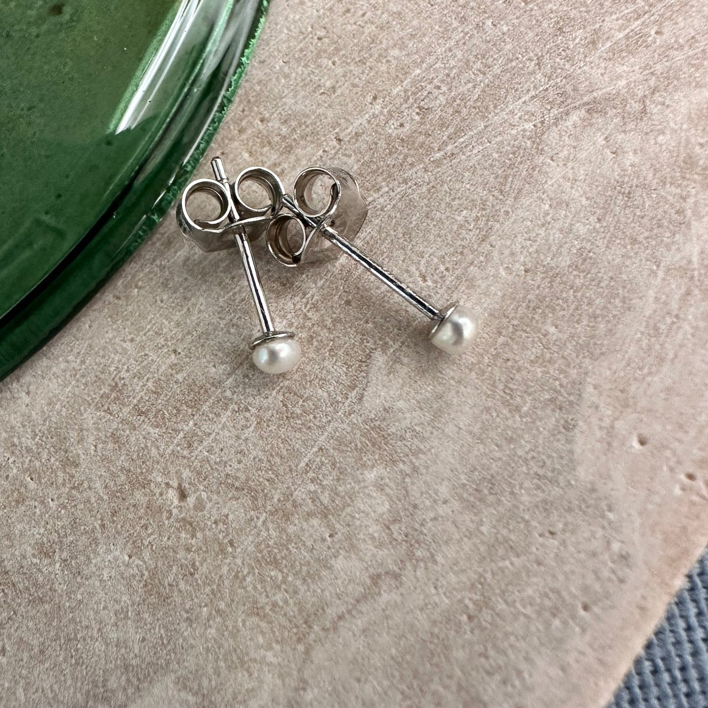 A pair of small-sized pearl stud earrings on a wooden board with a green glass object in the background.