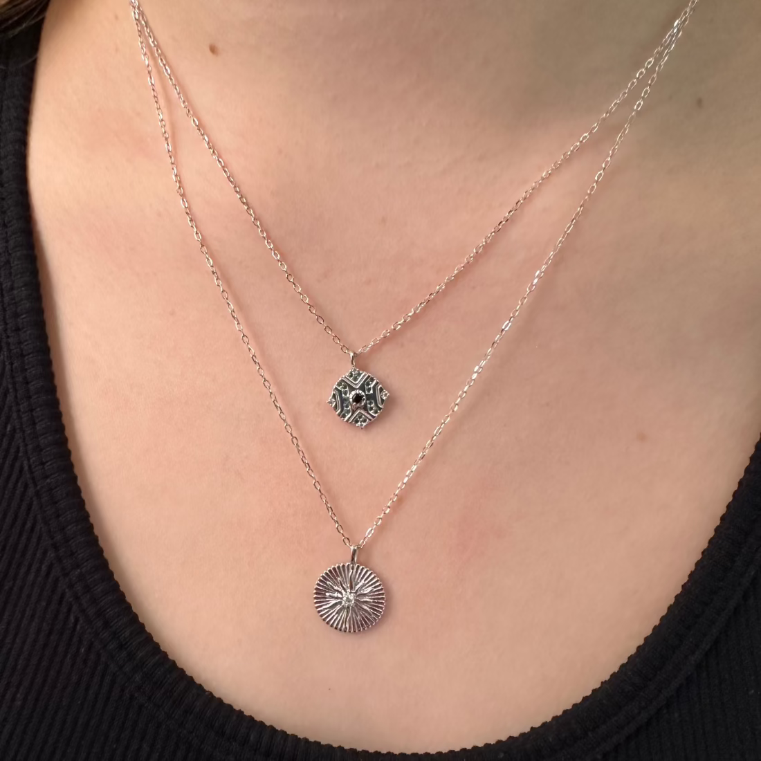a close up of models neck wearing two silver pendants of a sunburst and the other is a geometric moroccan inspired pendant