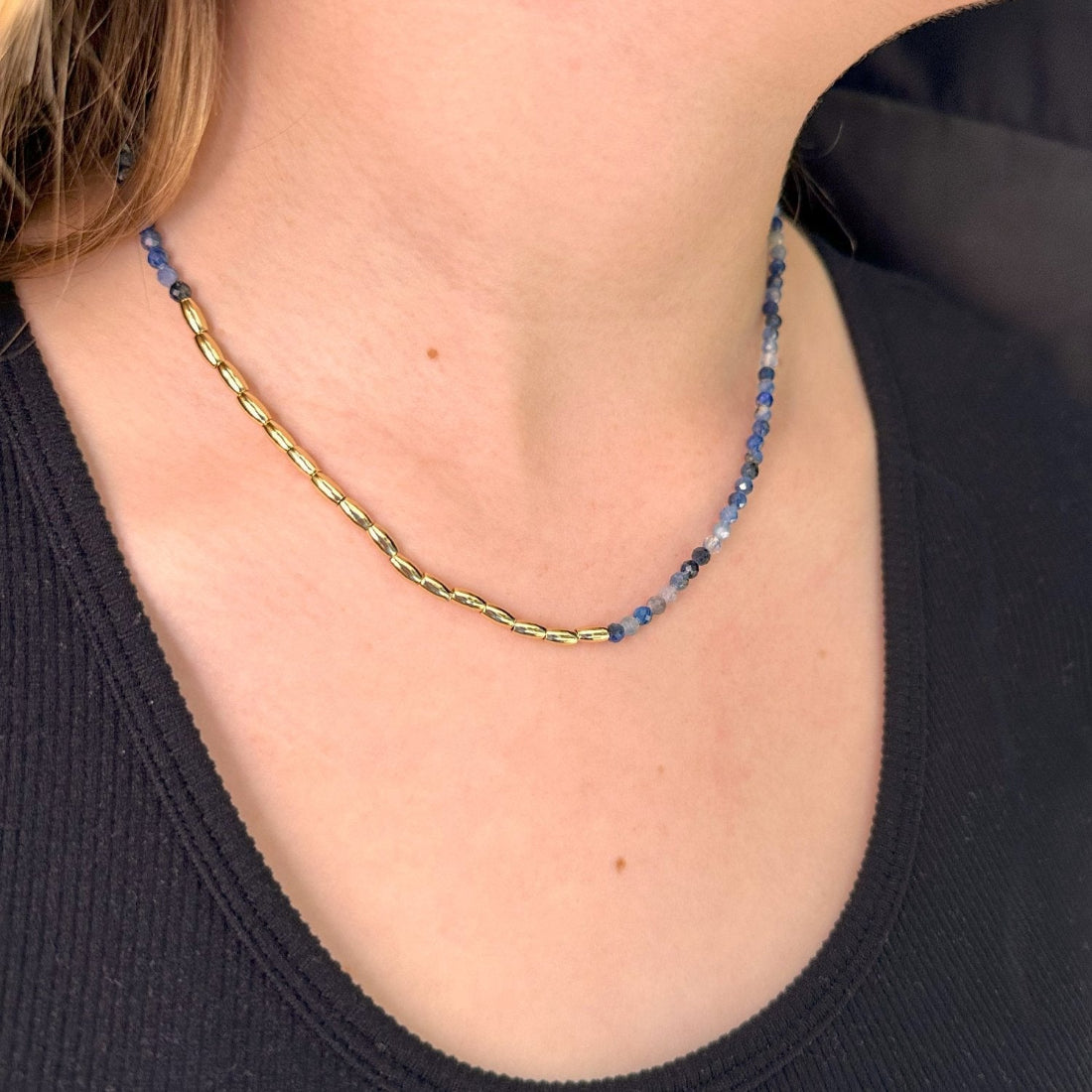 A close-up image of a person wearing a gold-colored asymmetric necklace with blue gemstone-shaped beads.