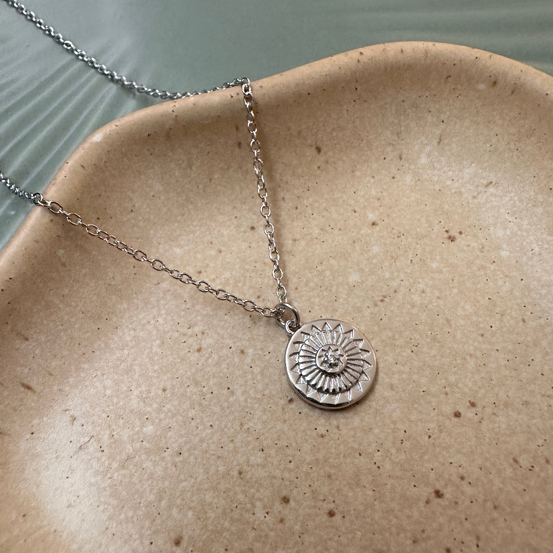 Silver geometric talisman necklace with a circular pendant on a beige ceramic plate