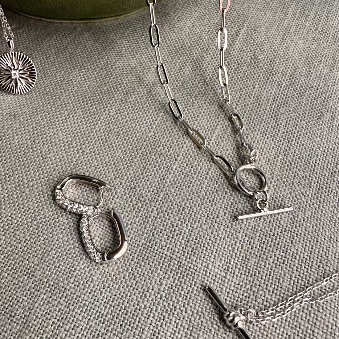 a silver T-bar toggle paperclip chain necklace with a sleek, modern design along with silver earrings with pave gemstones laid on greay material